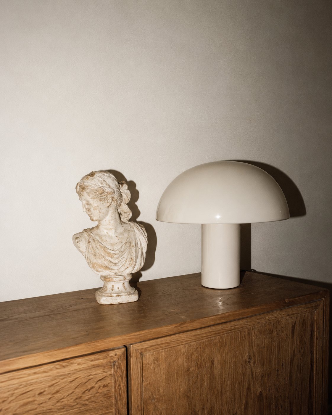 A weathered classical bust beside a cream mushroom lamp on an oak sideboard