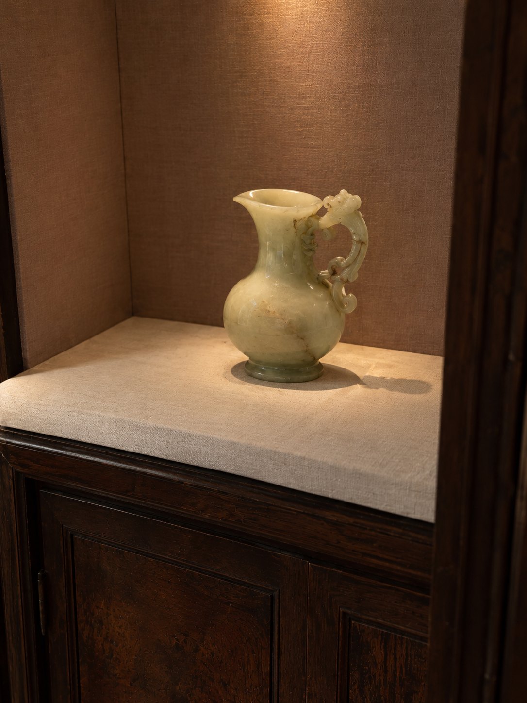 An antique pale jade ewer with a carved handle, lit in a linen-lined alcove above a dark wood cabinet